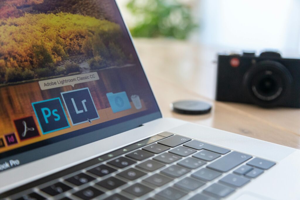 alt="Using Canva design tool as a beginner" Close-up of a laptop displaying Adobe software icons, beside a camera on a desk.