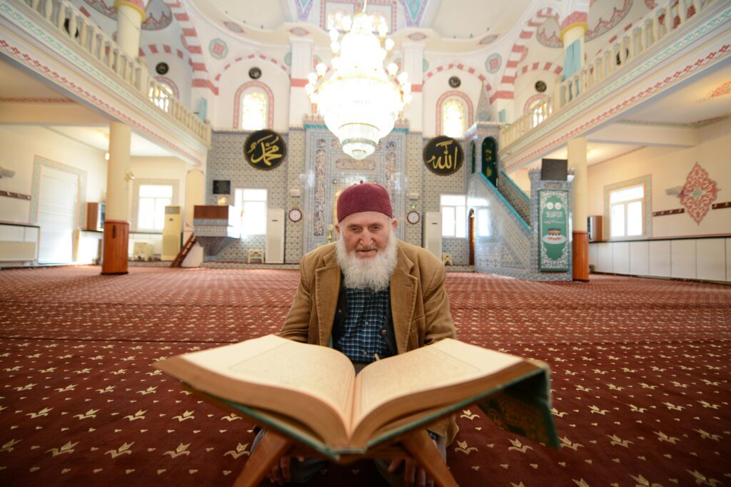 An elderly man with a beard reads the Quran inside a beautifully decorated mosque, emphasizing religious devotion.