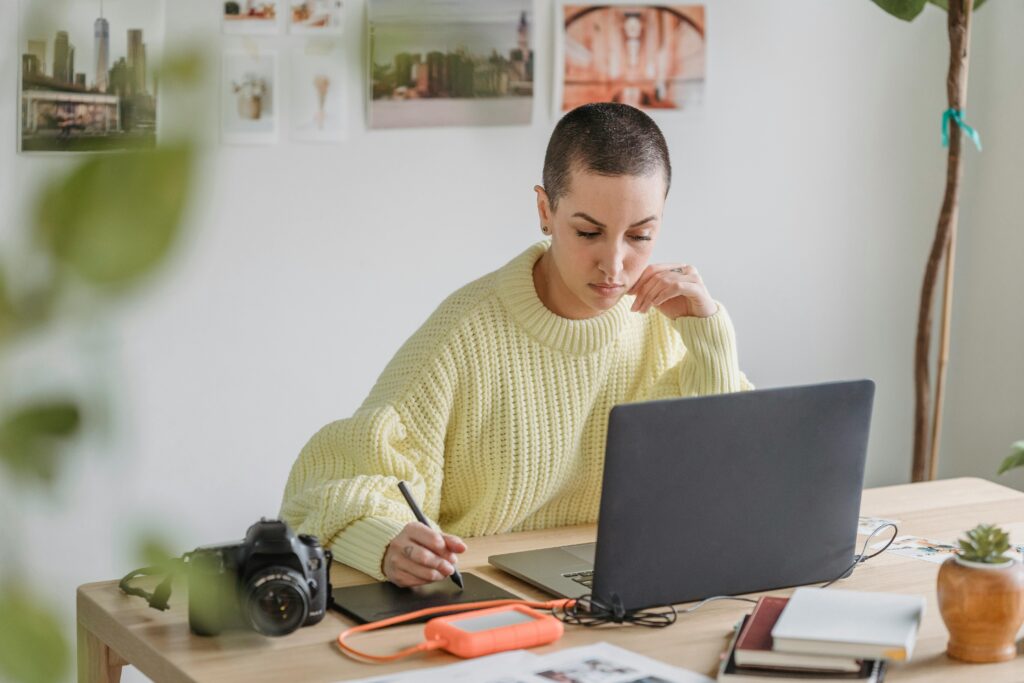 pexels-photo-7015100-7015100 Focused young female in casual sweater working on modern netbook and drawing on graphic tablet while sitting at desk