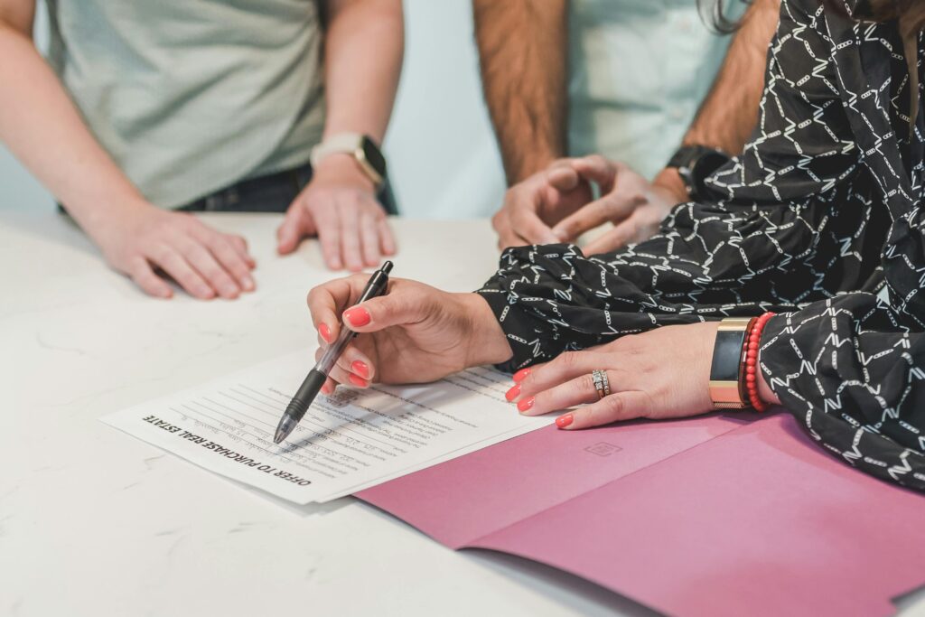 Close-up of hands signing a real estate document at a meeting table with three people.