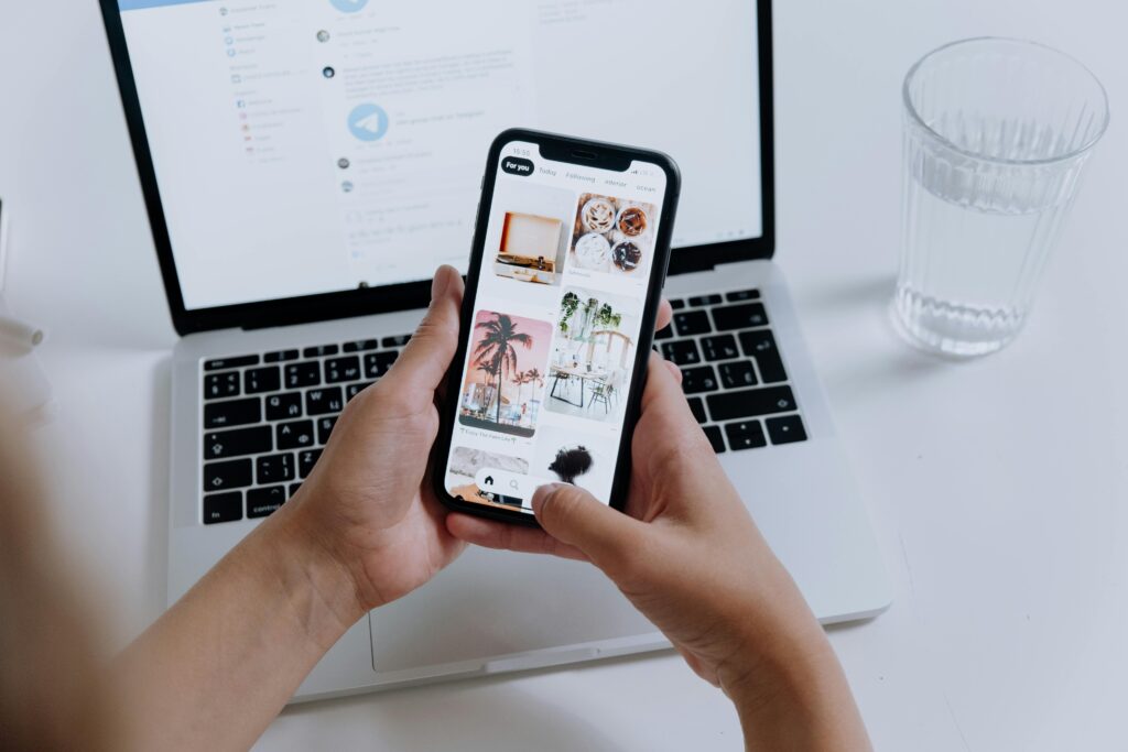pexels photo 5077039 5077039 Hands holding a smartphone showcasing a gallery, with a laptop in the background and a glass of water nearby.